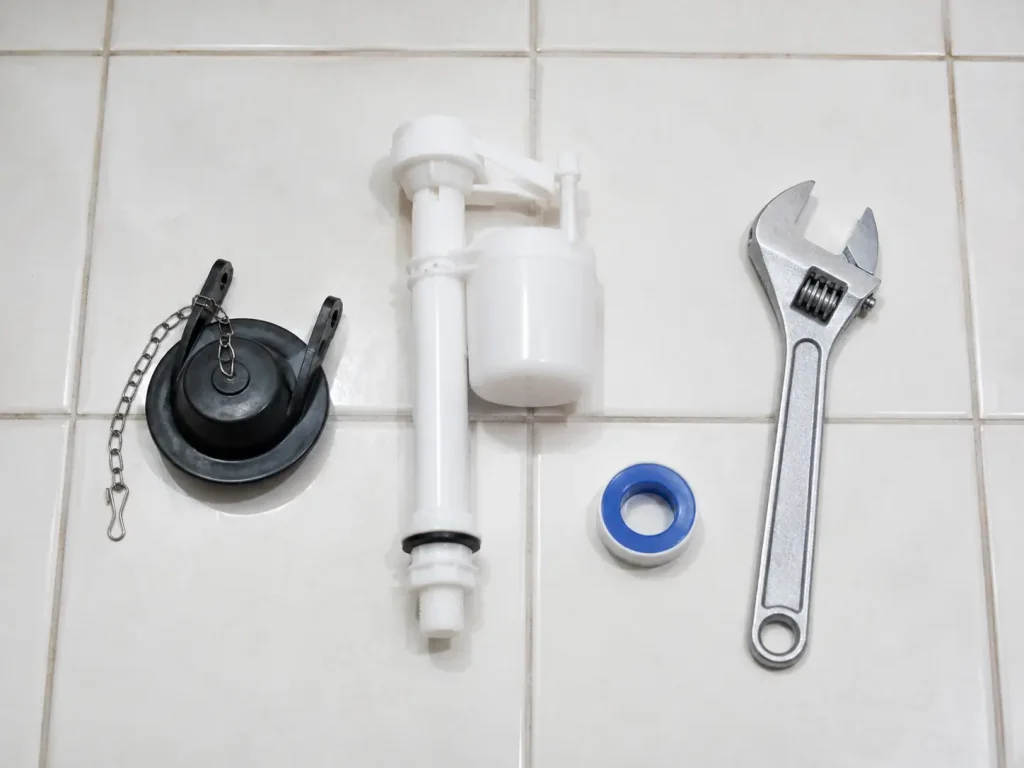Toilet repair parts and tools laid out on a tile floor including a flapper, fill valve, wrench, and thread tape