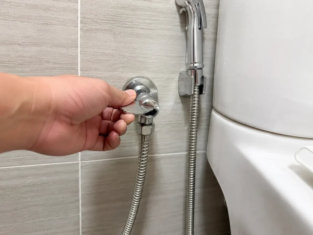 Hand turning off the wall angle valve before repairing a bidet spray hose.